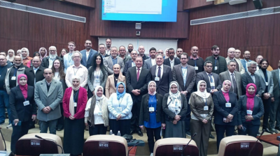 A large group of professionals, both men and women, pose together for a group photo in a conference room with a large screen behind them.