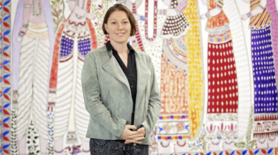 A woman stands indoors in front of a colorful, patterned mural featuring stylized figures in traditional attire.