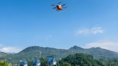 An orange drone with multiple rotors flies over a grassy field with distant hills and blue sky in the background.
