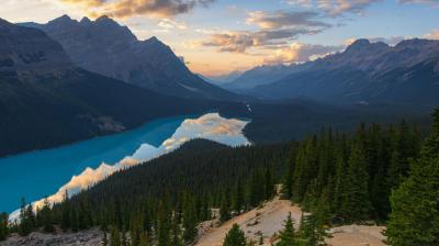 A turquoise lake winds through a forested valley surrounded by mountains at sunset, with clouds and sky reflected on the water's surface.