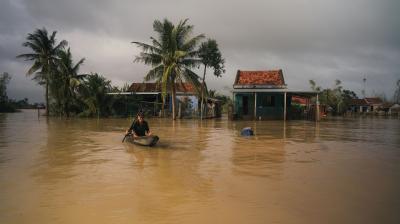 A person paddles a small boat through floodwaters in front of partially submerged homes and palm trees under a cloudy sky.