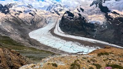 A wide glacier winds through a rocky mountain valley under a partly cloudy sky, with patches of snow on the surrounding peaks.