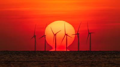 Wind turbines are silhouetted against a large setting sun over the ocean, with the sky glowing in shades of orange and red.