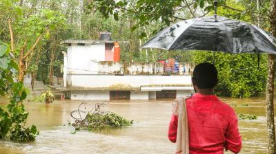 A person holding an umbrella observes a partially submerged house surrounded by floodwater and trees.