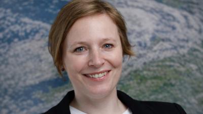 A woman with short light brown hair smiles at the camera, wearing a black blazer and white shirt, with a blurred Earth image in the background.