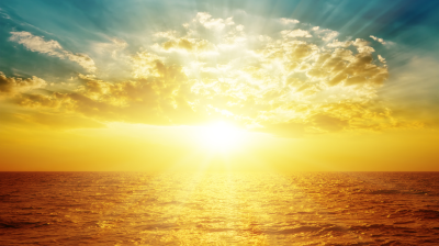 A bright golden sun rises or sets over the ocean, illuminating clouds and reflecting light on the water under a partly cloudy sky.