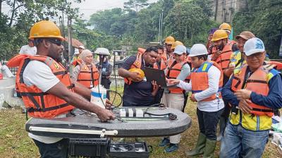A group of people wearing safety helmets and life vests gather outdoors around equipment, with one person using a laptop and another handling a survey instrument.
