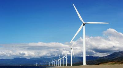 A row of wind turbines stands along a sandy coastline with mountains and blue sky in the background.