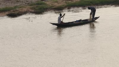Two people in a wooden boat are fishing with a net on a calm river near a grassy and sandy riverbank.