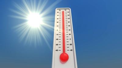A thermometer showing a high temperature stands against a clear blue sky with the sun shining brightly in the background.