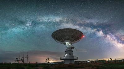 A large radio telescope dish stands under a star-filled night sky with the Milky Way galaxy visible overhead.