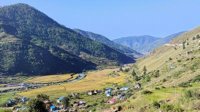 A river flows through a valley with yellow fields, small houses, and green hills under a clear blue sky.