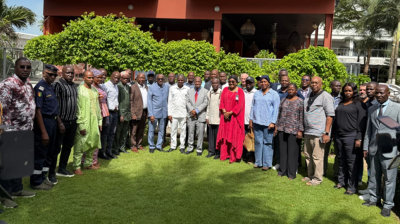 A large group of adults stands together outside on a lawn in front of a building and greenery, posing for a group photo in daylight.