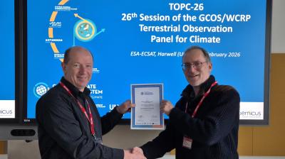 Two men shaking hands, one holding a certificate, in front of a screen displaying information about the 26th Session of the GCOS/WCRP Terrestrial Observation Panel for Climate.