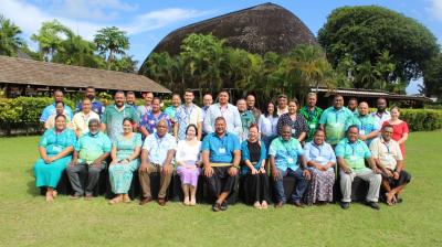 A large group of adults poses for a photo on grass in front of a tropical building with thatched roof and palm trees, under a partly cloudy sky.