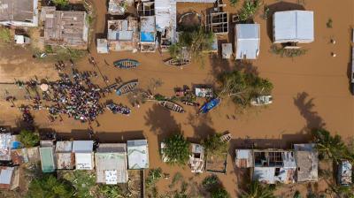 Aerial view of a flooded village with submerged houses, boats, and a large group of people gathered on a dry patch of ground.