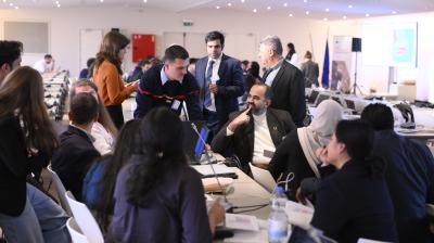 A group of people in business attire are gathered around tables with laptops, engaged in discussion at a conference or workshop in a well-lit room.