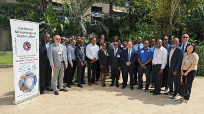 A group of people in formal attire stands outdoors next to a banner for the Caribbean Meteorological Organization. Trees and buildings are visible in the background.