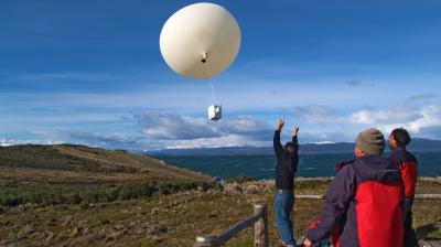 Three people on a wooden platform release a large weather balloon with attached instruments into a clear blue sky over a hilly, grassy landscape.