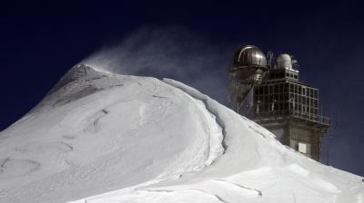 Snow-covered peak with wind-blown snow in the foreground and a building with a dome observatory structure in the background against a dark sky.