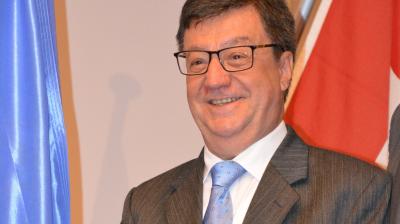 A man in a suit and glasses smiles while standing in front of blue and red flags.