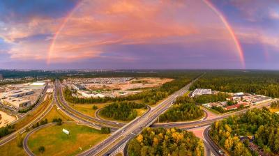 Aerial view of a highway interchange surrounded by greenery with a vivid rainbow arching across a partly cloudy sky at sunset.