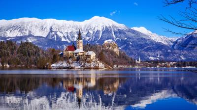A church and castle stand on Lake Bled’s island, surrounded by snow-capped mountains and reflected in the calm water under a clear blue sky.