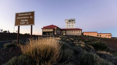A large building complex sits on a rocky hillside at sunset, with a sign reading "Observatorio Atmosférico de Izaña 2367 m" in the foreground.