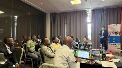 A group of people seated at tables in a conference room listen to a man presenting near a screen and a display banner. Laptops and bottles are on the tables.