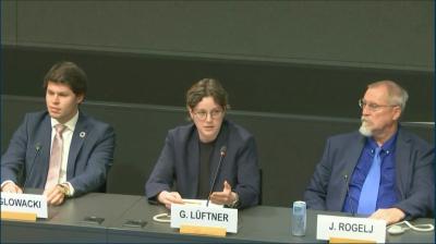 Three people sit at a panel table, with microphones and nameplates in front of them. The person in the center is speaking while the others listen.