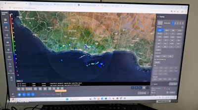 A computer monitor displays a weather radar interface showing precipitation data over a coastal region, with various control panels and map layers visible on the screen.