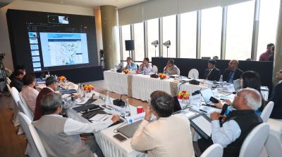A group of people in formal attire sit around a U-shaped table with laptops and documents, viewing a large screen displaying a map and data in a modern conference room.