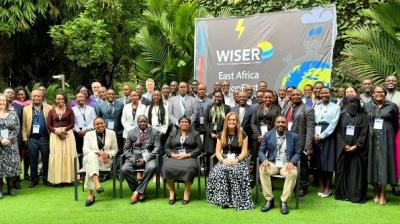 A large group of people pose for a photo outdoors in front of a sign that reads "WISER East Africa," surrounded by greenery.