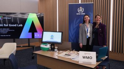 Two women stand smiling behind a desk with a "WMO" sign, in an office with a large screen displaying "AI for Good Lab" and WMO logo on the wall.