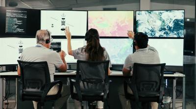 Three people wearing headsets sit at desks, facing large monitors displaying maps, satellite images, and data visualizations in a modern control room.