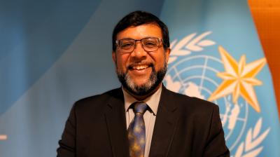 A man in a suit and tie smiles in front of a blue background featuring a United Nations-style emblem with a globe, laurel branches, and a star.