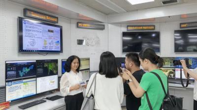 A woman stands in front of several monitors displaying weather data as she is interviewed and photographed by a group of people in a control room.
