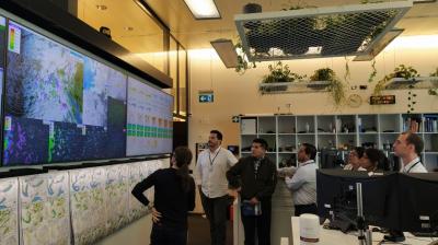 A group of people stand in front of large screens displaying weather maps and data in a modern office with plants and shelves in the background.