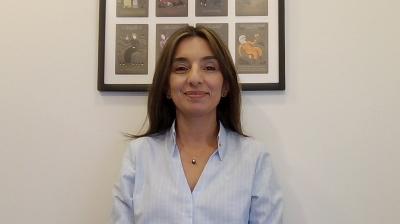 A woman with straight brown hair wearing a light blue shirt sits in front of a white wall with a framed artwork featuring several illustrations.
