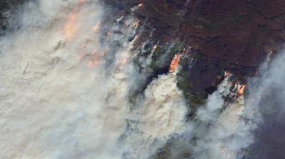 A large wildfire burns through a forested area, producing thick plumes of smoke and visible flames, as seen from an aerial or satellite perspective.