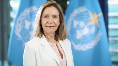 A woman in a white blazer stands in front of two blue flags featuring the United Nations emblem.
