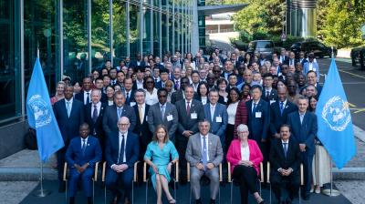 A large group of formally dressed people pose for a group photo outside a modern building, flanked by two United Nations flags.