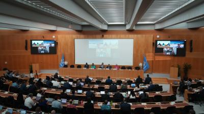 A conference room with attendees seated at desks facing a panel of speakers and a large screen, with United Nations flags displayed on both sides of the stage.
