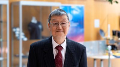 An elderly man in a dark pinstripe suit, white shirt, and red tie stands indoors with display cases and framed pictures in the background.