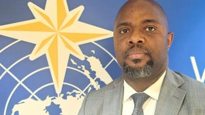 A man in a gray suit and tie stands in front of a blue wall featuring a yellow star and a world map design.