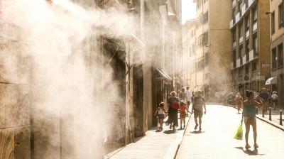 People walk down a sunlit street as mist or steam sprays from a building on the left, creating a hazy effect in the air.