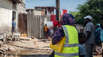 A person in a yellow vest holds a megaphone and documents while addressing people in an informal settlement area with scattered debris and makeshift structures.