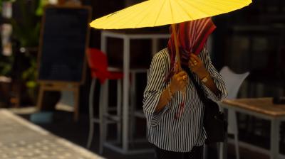 Person holding a large yellow umbrella, face covered by a striped scarf, standing on a sunlit sidewalk near outdoor seating and a blackboard sign.