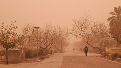 People walk along a tree-lined path under a heavy orange haze, likely caused by dust or wildfire smoke, limiting visibility and casting an unusual light over the scene.
