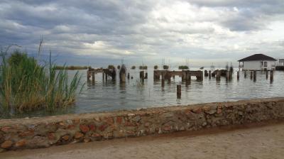 Sandy path with a stone wall, water with old wooden pilings, reeds on the left, and a small white building on the right under a cloudy sky.